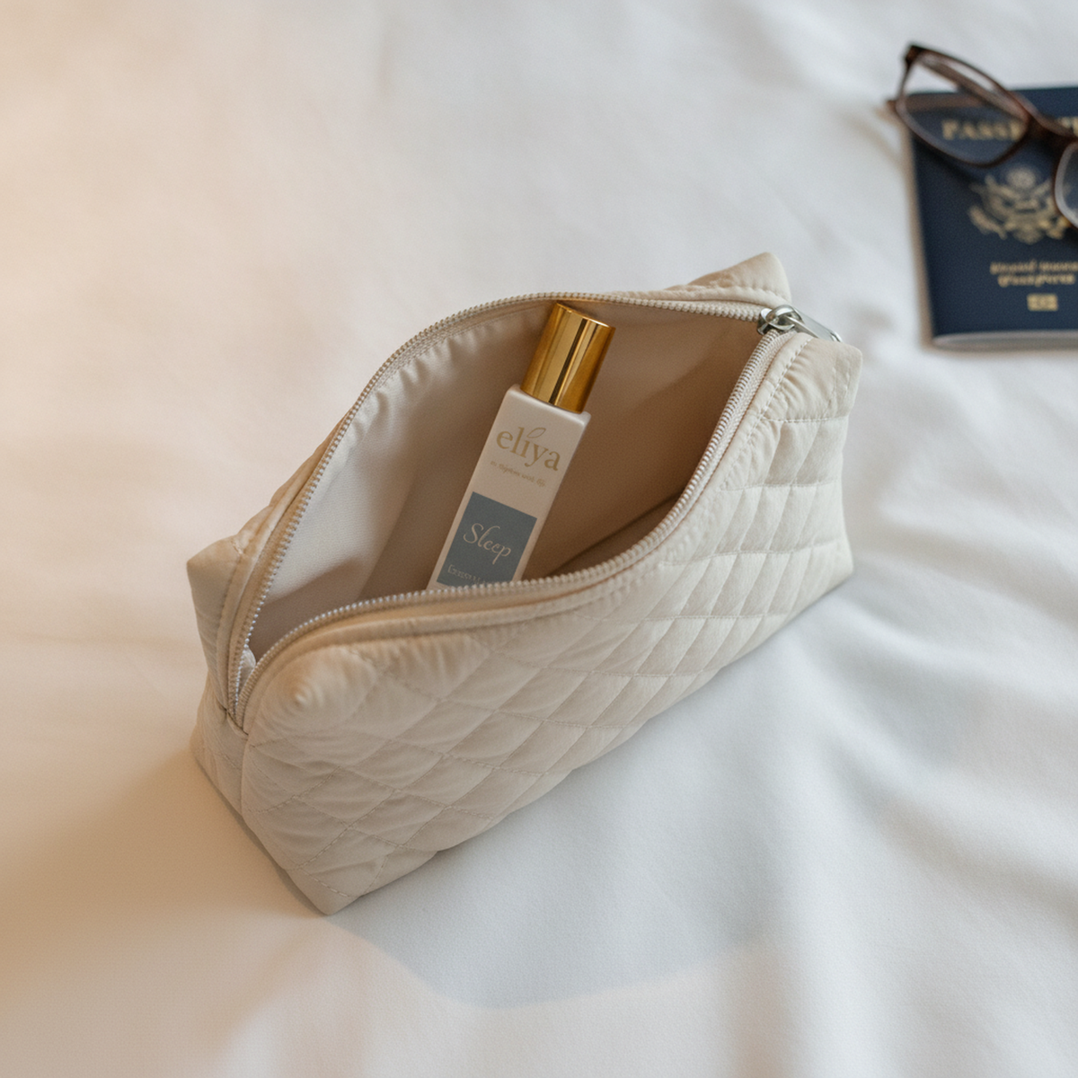 eliya Sleep roller standing inside a half-unzipped quilted travel pouch on a hotel duvet beside a passport and folded reading glasses