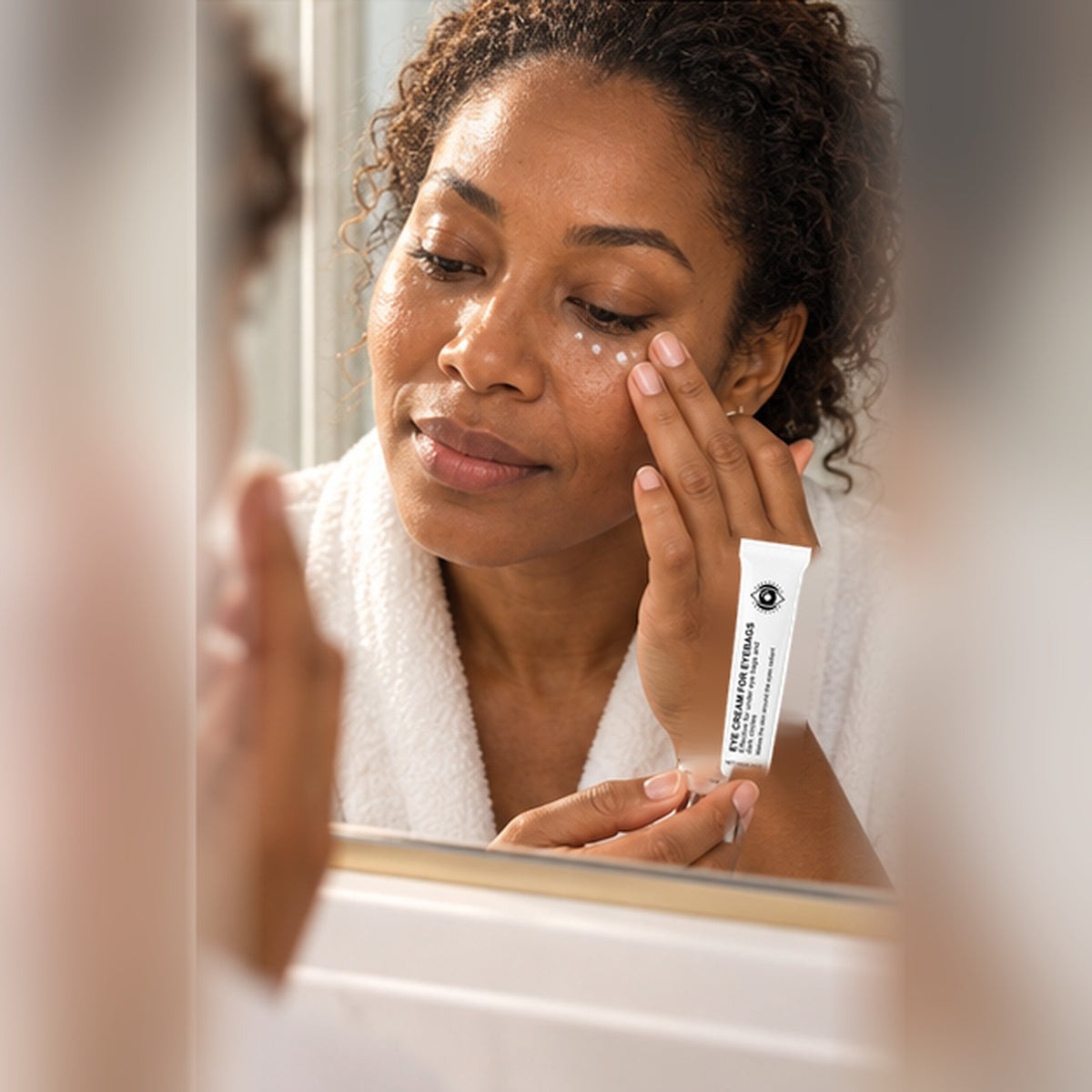 Woman at a bathroom mirror patting the eye cream under her eye, tube in hand