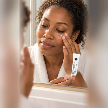 Woman at a bathroom mirror patting the eye cream under her eye, tube in hand